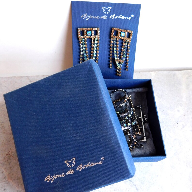 May include: A blue box with the text "Bijoux de Boh&egrave;me" and a butterfly logo. The box contains a pair of dangle earrings with a gold frame and blue and clear crystals. The earrings are in a clear plastic bag.