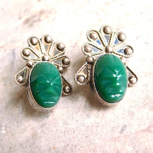 以下が含まれることがあります： 楕円形に彫られた緑色の宝石をあしらった、銀色のイヤリングのペア。イヤリングは、宝石の上に装飾的な銀色のデザインがあり、詳細で質感のある外観です。イヤリングは明るい色の表面に表示されています。
