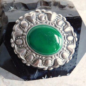 May include: A silver brooch with a green cabochon stone set in a detailed, ornate design.