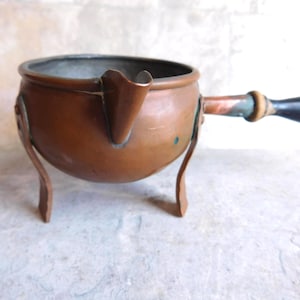 May include: A round copper pot with a pouring spout, supported by three curved metal legs. The pot features a long handle with a dark wooden grip and a metallic ferrule. The interior of the pot has a silver-toned lining, and the exterior shows a warm, metallic copper finish with subtle signs of age and wear.
