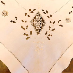 May include: Close-up of a white, intricately embroidered tablecloth. The design features a central diamond-shaped openwork pattern, surrounded by leaf-like cutouts and delicate floral accents. The edges are scalloped and finished with a stitched border.
