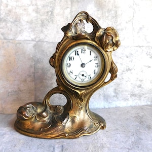 May include: A gold-toned metal mantel clock featuring an asymmetrical, flowing frame decorated with sculpted rose blossoms and leaves. The circular clock face has a white background with black Arabic numerals and two thin black hands.