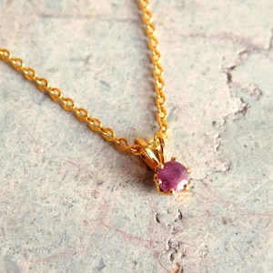 May include: A delicate gold-colored necklace featuring a small, round, ruby-colored gemstone pendant. The pendant is held in a gold setting and hangs from a fine chain. The necklace is displayed on a textured, light gray surface.
