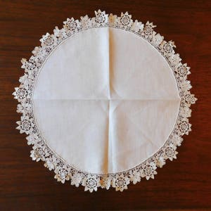 May include: White crocheted doily with a delicate floral pattern. The doily is round and has a scalloped edge.