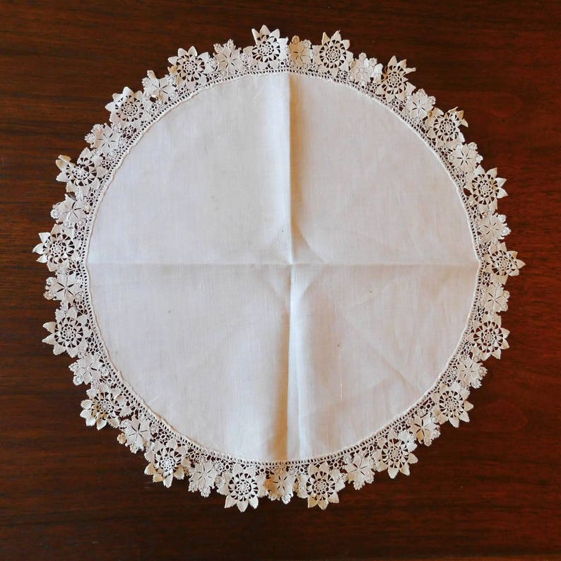 May include: White crocheted doily with a delicate floral pattern. The doily is round and has a scalloped edge.