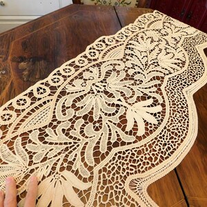 K&ouml;nnte beinhalten: Eine zarte cremefarbene Spitzenborte mit floralem Muster. Die Spitze ist filigran und detailliert mit einem gewellten Rand.