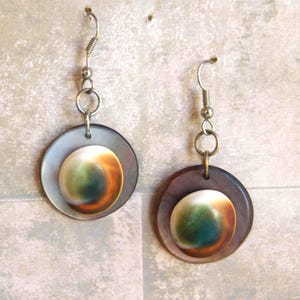 Pode incluir: Um par de brincos pendurados. Cada brinco apresenta um disco exterior circular, iridescente, cinza-marrom com um disco interior menor, redondo e multicolorido. Os discos interiores têm uma mistura de tons brancos, verdes e laranja. Os brincos têm ganchos prateados.