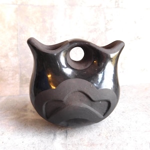 May include: A black ceramic bowl with a glossy finish. The bowl has a unique shape with a circular opening and a textured, wave-like design around the base. The bowl is approximately 6 inches in diameter.