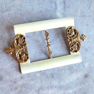 Könnte beinhalten: Eine rechteckige Gürtelschnalle mit einem glatten, cremeweißen Rahmen und verzierten, goldfarbenen Metallfiligranen an der linken und rechten Seite. Ein zentraler vertikaler Metallsteg mit kleinen floralen Details verbindet den oberen und unteren Rahmen.