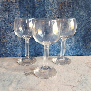 May include: Three clear glass wine glasses with a fluted stem and a round bowl. The glasses are arranged in a triangle shape on a light gray surface.