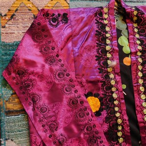 K&ouml;nnte beinhalten: Ein magentafarbenes, satinartiges Kleidungsst&uuml;ck mit aufwendigen, schwarz gestickten Blumenmustern. Das Kleidungsst&uuml;ck hat eine schwarze und goldene Bord&uuml;re und ein gelbes Blumendetail. Das Innenfutter hat einen schwarzen Hintergrund mit bunten Punkten.