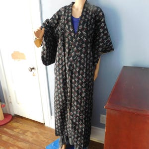 May include: A long, black kimono with a repeating floral pattern in white and red. The kimono is displayed on a mannequin, showcasing its full length and wide sleeves. The garment is open in the front, revealing a blue top underneath.