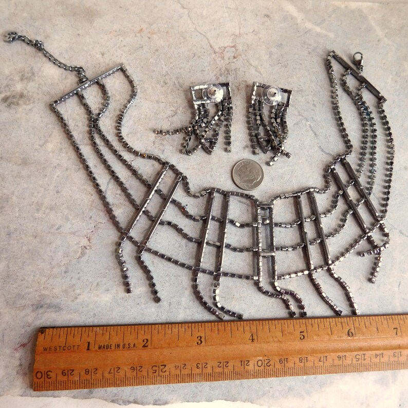 May include: A silver tone rhinestone choker necklace and matching earrings. The necklace is a geometric design with a fringe of rhinestones. The earrings are also geometric with a fringe of rhinestones.