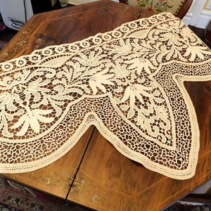K&ouml;nnte beinhalten: Eine zarte cremefarbene Spitzentischdecke mit einem gewellten Rand und komplizierten Blumenmustern. Die Spitze ist zart und filigran, mit einer feinen Webart und einer weichen Textur.