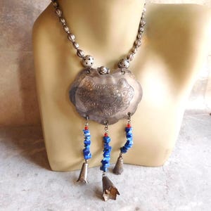 May include: A silver-toned necklace featuring a large, ornate, shield-shaped pendant with intricate engraved patterns. Three dangling strands hang from the bottom, each composed of small blue stone chips, red beads, and a small silver-toned bell.