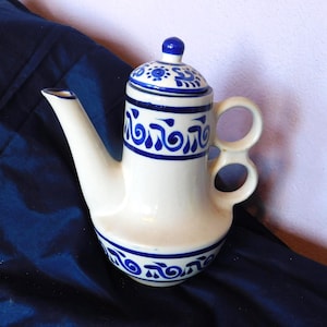 May include: A cream-colored ceramic teapot featuring hand-painted cobalt blue scrollwork patterns around the lid, neck, and base. The teapot has a long, tapered spout and a unique handle design consisting of two stacked circular loops.