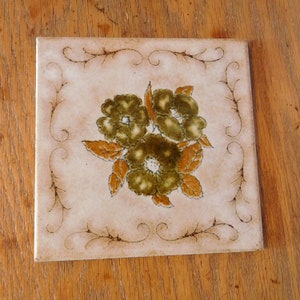 May include: A white ceramic tile with a brown floral design. The flowers are green and brown with a white center. The tile has a decorative border of brown swirls.