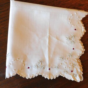 May include: A white linen handkerchief with a scalloped edge and embroidered with a delicate floral pattern in shades of green and red.
