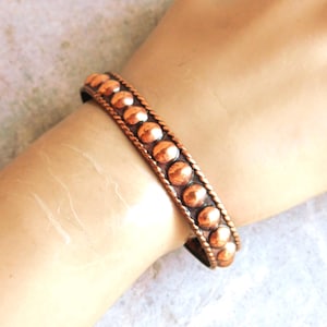 May include: A copper-toned bracelet with a series of rounded studs. The bracelet has a braided border on each side. The bracelet is approximately 0.75 inches wide.