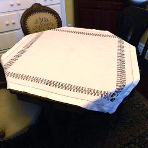 May include: A white octagonal tablecloth with a detailed black embroidered border. The tablecloth is draped over a dark wooden table, showcasing its elegant design. The embroidered border features a repeating geometric pattern.