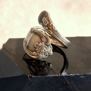 May include: A silver ring, crafted from repurposed silverware, with floral detailing. The ring's design incorporates the spoon bowl and handle. The ring is displayed on a black surface, highlighting its intricate design and vintage appeal.