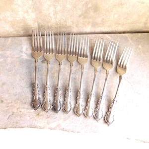 Puede incluir: Vista aérea de ocho tenedores antiguos de plata. Cada tenedor tiene un mango largo y delgado con detalles ornamentados y una cabeza de cuatro púas. Los tenedores están dispuestos sobre una superficie clara y texturizada, sugiriendo una colección vintage.