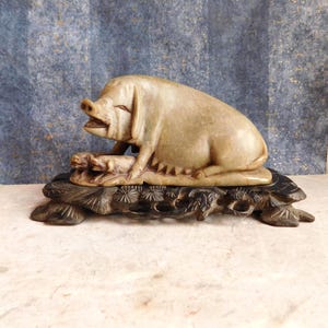 May include: A carved stone sculpture of a pig lying on a carved wooden base. The pig is light beige with detailed features. The base is dark brown with intricate floral carvings. The sculpture is a decorative item.