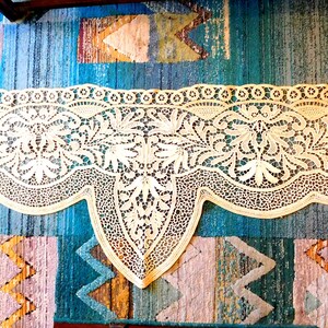 K&ouml;nnte beinhalten: Eine zarte, cremefarbene Spitzenborte mit einem gewellten Rand und filigranen Blumenmustern. Die Spitze liegt auf einem bunten gemusterten Teppich.