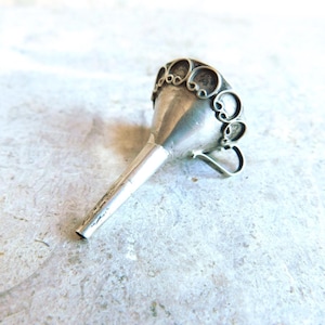 Puede incluir: Un pequeño embudo plateado con un borde calado decorativo. El embudo tiene un pico largo y cónico y un pequeño asa. El borde presenta diseños circulares y en forma de volutas. El objeto es probablemente de metal.