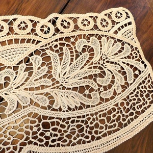 K&ouml;nnte beinhalten: Eine zarte cremefarbene Spitzentischdecke mit floralem Muster. Die Tischdecke hat einen gewellten Rand und ist aus feiner, filigraner Spitze gefertigt.