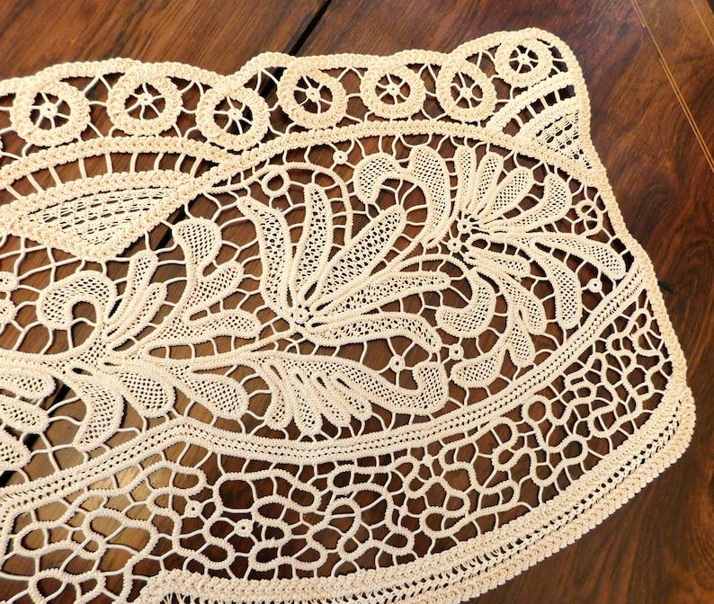 K&ouml;nnte beinhalten: Eine zarte cremefarbene Spitzentischdecke mit floralem Muster. Die Tischdecke hat einen gewellten Rand und ist aus feiner, filigraner Spitze gefertigt.