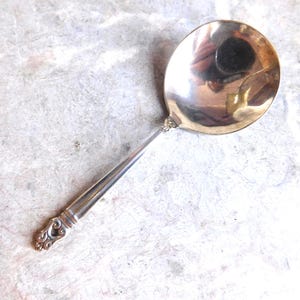 May include: A polished silver-toned serving spoon with a wide, rounded bowl and a slender handle. The handle features a decorative, ornate scrollwork design at the base, while the bowl has a reflective surface showing a distorted reflection.