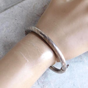 May include: A silver-toned hinged bangle bracelet featuring a polished finish with a series of diagonal etched lines running across the surface. The bracelet has a secure clasp mechanism and a rounded, tubular profile that encircles the wrist.