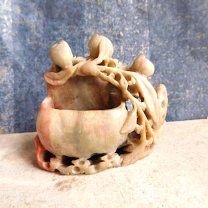 May include: A carved stone sculpture featuring a bowl-like shape with intricate floral and leaf details. The sculpture has a natural color palette of beige, pink, and red hues. The piece is likely a decorative item or a small container.