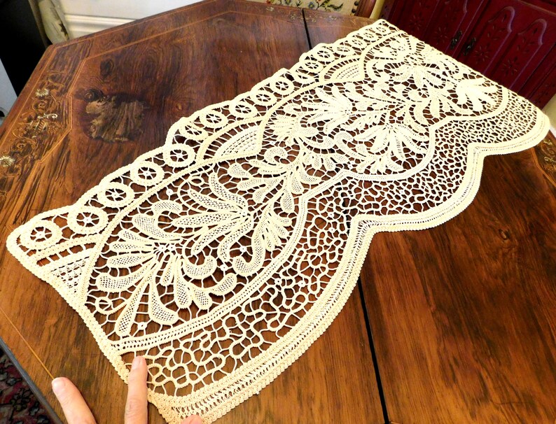 K&ouml;nnte beinhalten: Ein zarter cremefarbener Tischl&auml;ufer aus Spitze mit einem gewellten Rand und filigranen Blumenmustern. Der L&auml;ufer liegt auf einem dunklen Holztisch.
