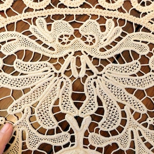 K&ouml;nnte beinhalten: Nahaufnahme einer zarten, cremefarbenen H&auml;keltischdecke mit filigranen Blumenmustern. Die Spitze ist in einem feinen, komplizierten Design gewebt, das einen zarten und eleganten Look erzeugt.