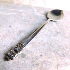 May include: A silver-toned metal spoon featuring a polished oval bowl and a long, slender handle. The handle is decorated with a detailed, raised scrollwork pattern at the base, consisting of symmetrical swirling motifs and horizontal ridges.