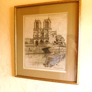 May include: A black and white etching of Notre Dame Cathedral in Paris, France. The cathedral is shown from a distance, with the Seine River in the foreground. The etching is framed in a gold-colored frame.