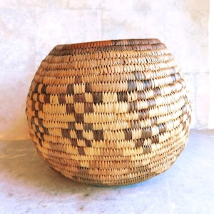 May include: A round woven basket with a natural color scheme. The basket features a repeating pattern of light and dark brown woven elements. The basket has a wide opening and a rounded shape, suitable for storage or display.