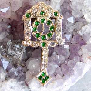 May include: A gold-toned brooch with a geometric design featuring a circle of green stones surrounded by smaller white stones. The brooch is shaped like a key and has a pointed end.