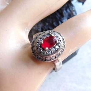 May include: A silver-toned ring featuring a central oval-shaped deep red gemstone surrounded by a halo of small, clear, sparkling stones. The setting is oval, and the band is a smooth, solid silver-colored metal that encircles the finger.