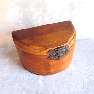 May include: A semi-circular wooden box with a smooth, light brown finish and a visible wood grain. The container features a hinged lid secured by an ornate, dark metal latch centered on the front edge, connecting the lid to the base.