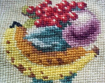Könnte beinhalten: Nahaufnahme eines Stickbildes mit einer Banane, einer lila und grünen Frucht und einer Traube roter Beeren.