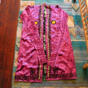 K&ouml;nnte beinhalten: Eine lange, flie&szlig;ende magentafarbene Robe mit aufwendiger schwarzer Blumenstickerei und gelben Blumenakzenten. Die Robe hat ein schwarzes und gemustertes Innenfutter mit gr&uuml;nen und gelben kreisf&ouml;rmigen Details. Das Kleidungsst&uuml;ck ist aus gl&auml;nzendem, seidigem Material gefertigt.