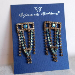 May include: A pair of gold-toned dangle earrings with a square-shaped top and a cascading fringe of iridescent blue and brown crystals. The earrings are displayed on a blue card with the text "Bijou de Boh&egrave;me".