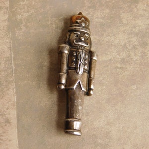 Pode incluir: Uma figura de quebra-nozes prateada com um rosto detalhado e um uniforme preto e branco. O quebra-nozes está em pé e tem uma pequena coroa na cabeça.