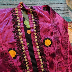 K&ouml;nnte beinhalten: Eine magentafarbene Robe mit aufwendiger schwarzer und goldener Stickerei. Die Robe hat ein Blumenmuster und einen kontrastierenden schwarzen Einsatz mit bunten Tupfen. Die Robe hat eine Tasche mit einem gelben Blumendesign.