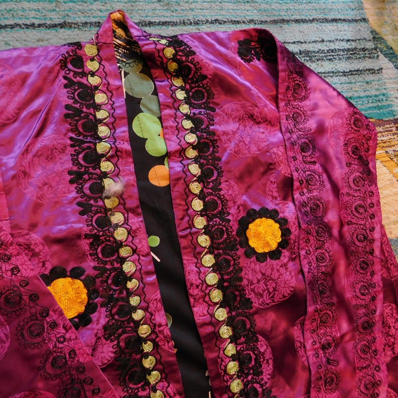 K&ouml;nnte beinhalten: Eine magentafarbene Robe mit aufwendiger schwarzer und goldener Stickerei. Die Robe hat ein Blumenmuster und einen kontrastierenden schwarzen Einsatz mit bunten Tupfen. Die Robe hat eine Tasche mit einem gelben Blumendesign.