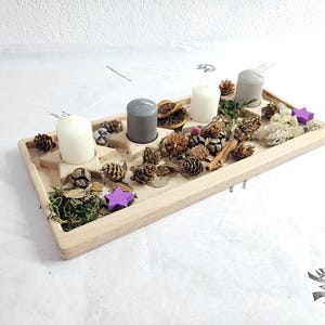 Puede incluir: Un portavelas de Adviento de madera con cuatro velas, dos blancas y dos grises. El portavelas está decorado con piñas, musgo, rodajas de naranja secas y adornos en forma de estrella morada. El diseño es rústico y natural.