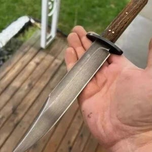May include: A handcrafted knife with a polished steel blade and a dark wooden handle. The handle is secured with small brass rivets. The knife has a black bolster and a sharp, pointed tip. The knife is held in a hand.